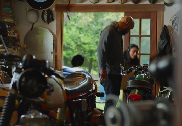A couple walks into a workshop from the yard outside holding a plastic bin and cardboard box and then touch and examine one of the motorcycles inside.