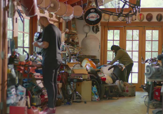 A bald, bearded man and young brunette woman rummage through the stuff in a cluttered garage and the woman finds a golf bag full of clubs hidden in the corner and drags it out, holding it up for him to see.