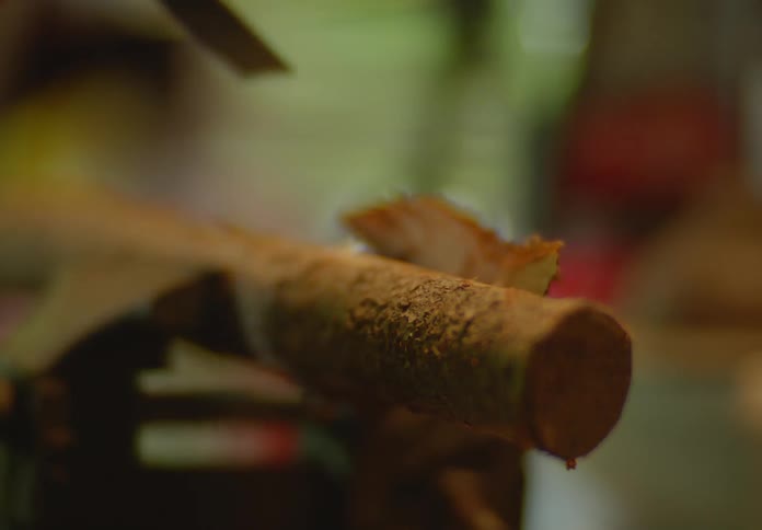 Hands of a male woodcarver using a metal chisel and hammer to remove bark from a small log in small shavings.