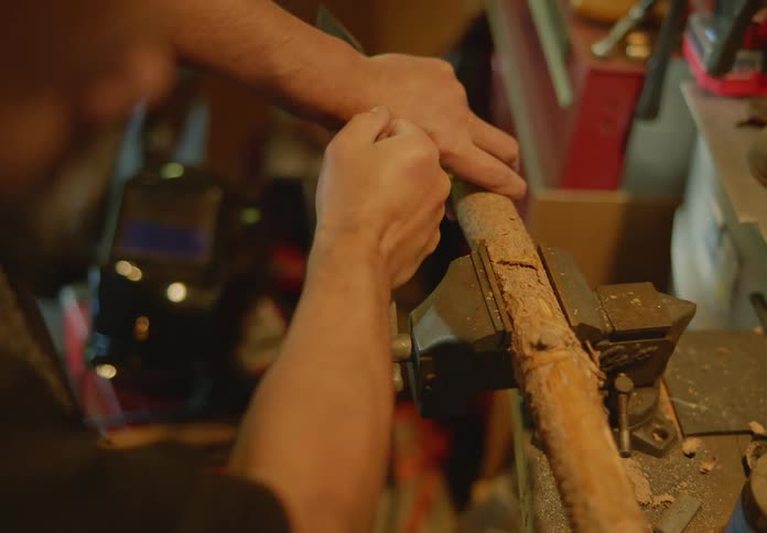 A bearded man uses the rotating handle on a vice to secure a piece of wood that he is about to carve with a hammer and chisel.