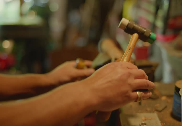 A man is using a hammer and chisel to carve into a small log held in a vice which slips, and the man adjusts it and starts again.