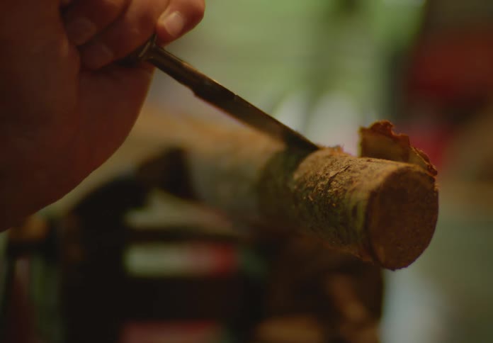 Close-up of a woodworker's hand using a metal chisel to shave off the bark layer of a small log held in a metal vice.
