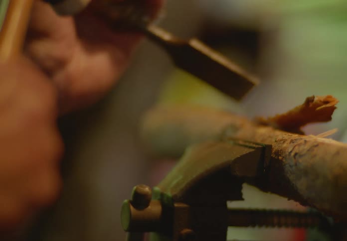 A bearded, bald man focuses intently on his hobby of woodcarving as he uses a hammer and chisel to carve bark shavings off of a small log being held in a metal vice.