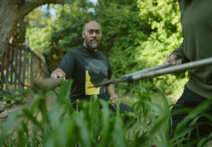 Man holds staff to help woman trim plants to the same height in their garden