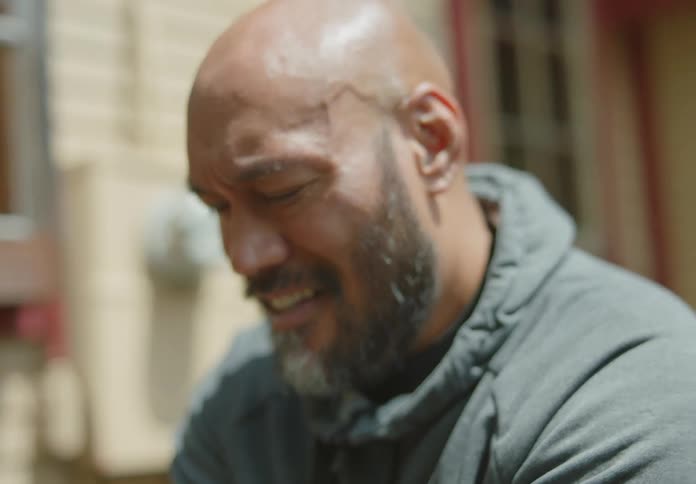 A fit and tan Pacific-Islander man sits on a motorcycle and adjusts something near the dashboard while laughing and talking with a young dark-haired woman beside him.