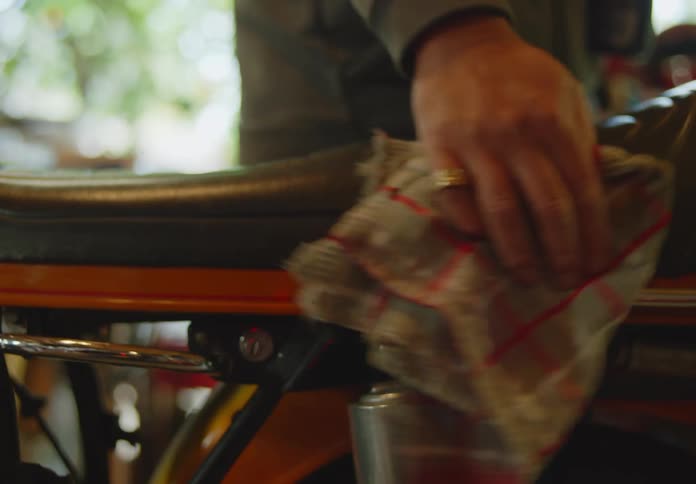 A man wearing a dark hoodie is polishing a motorcycle seat with a rag while holding a spray bottle.