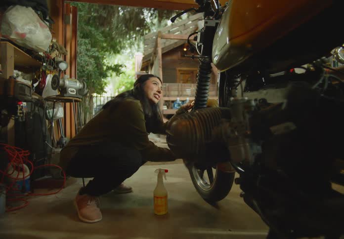 A young, dark-haired woman is squatting down and cleaning the tire of a motorbike in an outdoor workshop with natural light