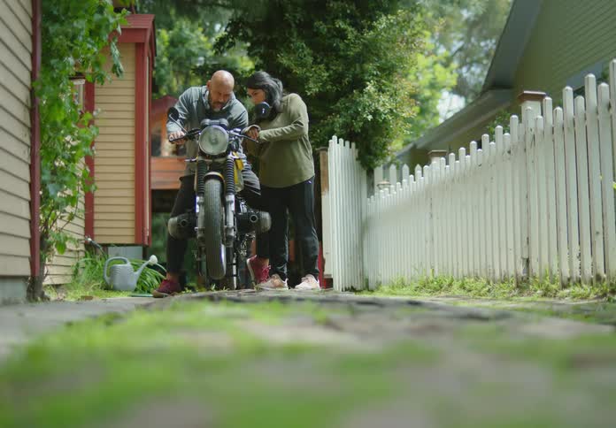 A bald, bearded man sitting on a motorcycle in a driveway shows a dark-haired young woman standing next to him how it operates