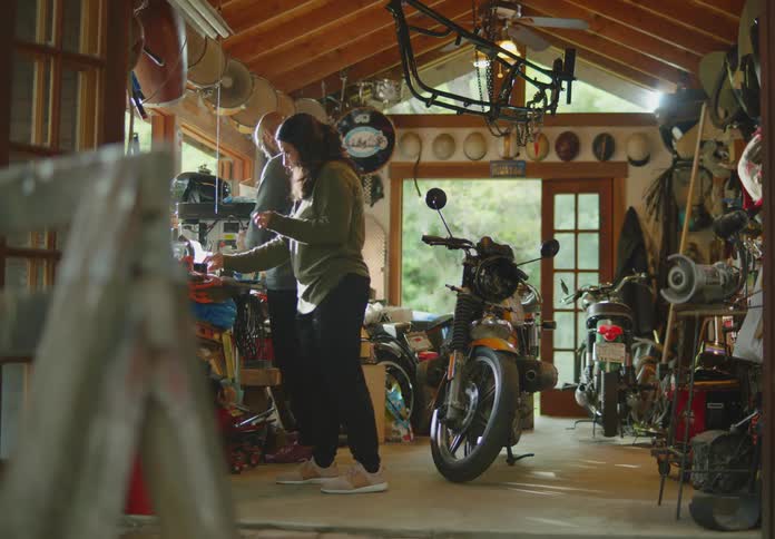 A man and a woman work on a hobby together in a cluttered garage workshop filled with motorcycles and lots of other items.