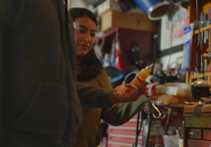 A man and a woman are in a garage workshop and the man passes her a small plastic bottle of an orange liquid as he walks by a motorcycle with no headlight
