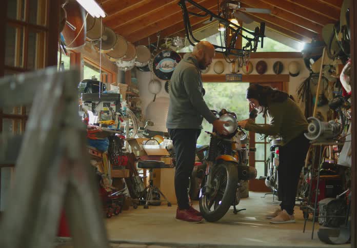 A young, ethnic woman standing in a messy workshop turns to grab a bottle of glue and then talks with the bald, bearded man standing with her as he demonstrates how they will install it.