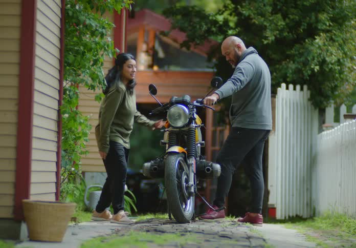 A bald, bearded man shows off his motorbike to a young, dark-haired female outdoors on an unpaved driveway