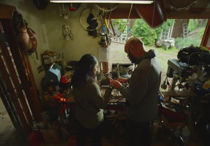A bearded and bald man wearing a hoodie and a woman with dark hair look at tools and parts on a workbench and discuss them.