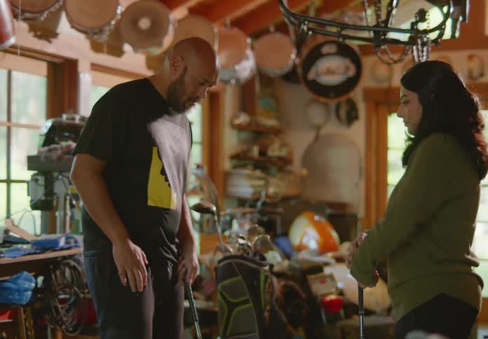 A bald, bearded man and a young brunette female holding golf clubs stand in a cluttered garage and the man shows her technique on how to stand when preparing to swing.