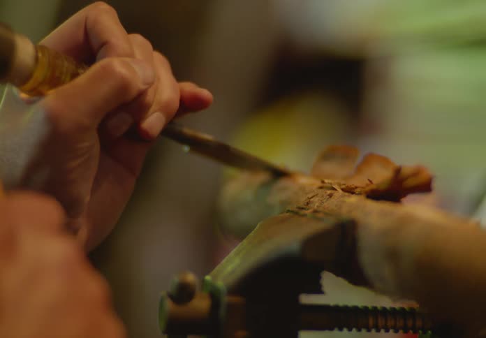 A woodcarver's hands using a hammer and chisel to carve shavings off of a piece of wood that is clamped in a vice.