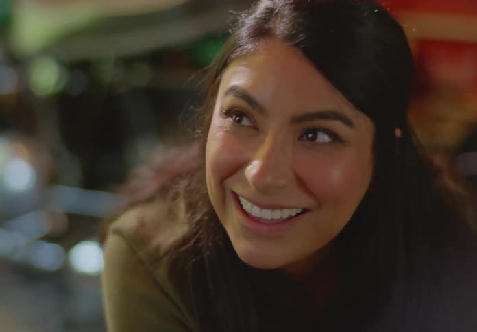 A young, dark haired ethnic woman smiles and makes a surprised facial expression then blows air into her cheeks and back out as she laughs and smiles at something she finds funny..