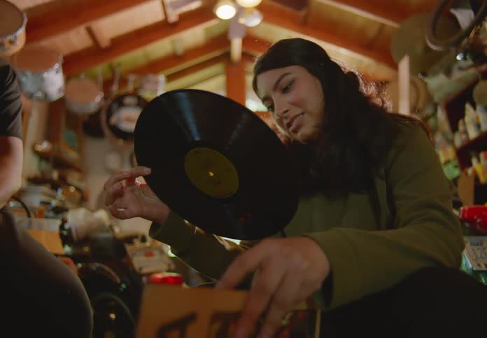 A dark-haired young woman crouching down in a crowded storage room pulls an old vinyl record out of a crate and flips it over and back while discussing it with the man seated next to her.