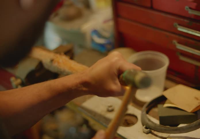 View over the shoulder of a man at his workbench with a hammer and chisel carving into a piece of wood being held in a metal vice.