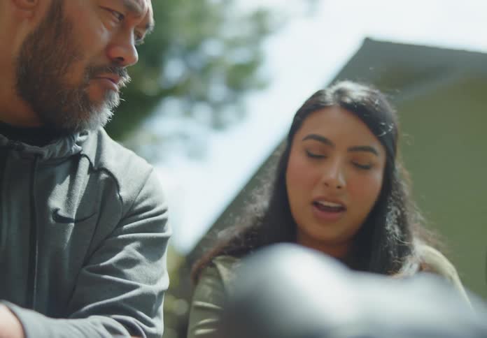 POV from below of a bald, bearded man in a hoodie sitting on a motorbike explaining something about it to a dark-haired young woman standing next to him as she laughs, gestures sarcastically, and smiles.