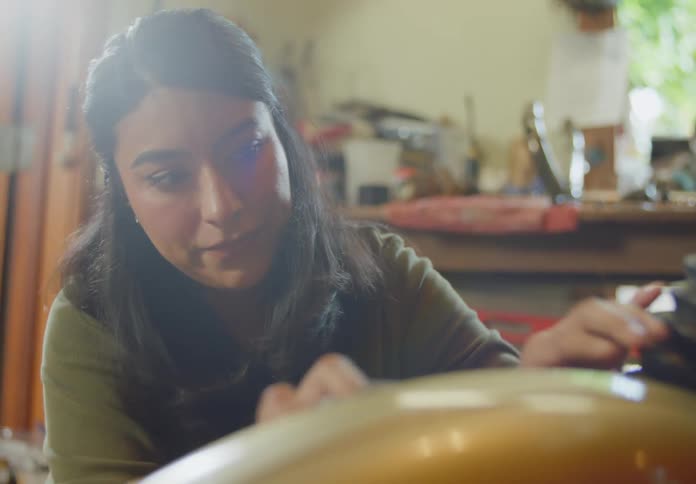 A dark-haired ethnic woman is bending over carefully polishing and detailing a motorcycle with a cloth