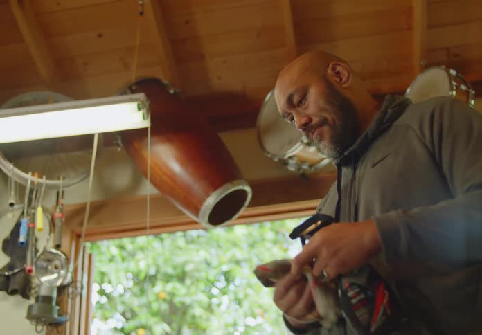 A bald man with beard and dark complexion stands in a workshop with drums hanging from the ceiling and uses a spray bottle and rag to wipe something down while talking to someone off screen.