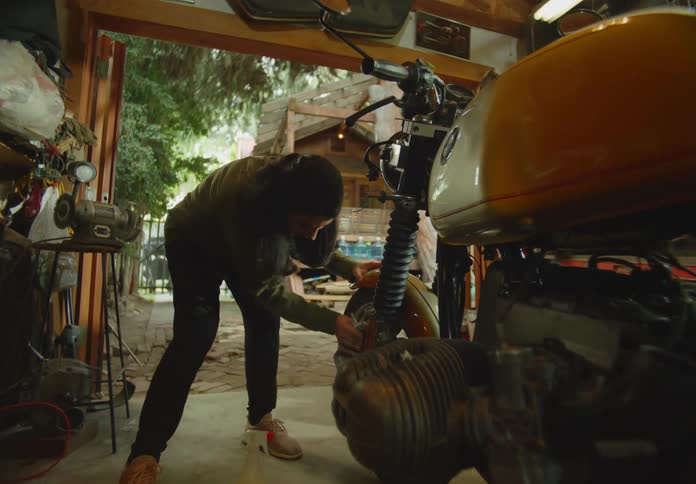 A young dark-haired woman is bending over in an open-air workshop and cleaning a motorcycle tire with a rag.