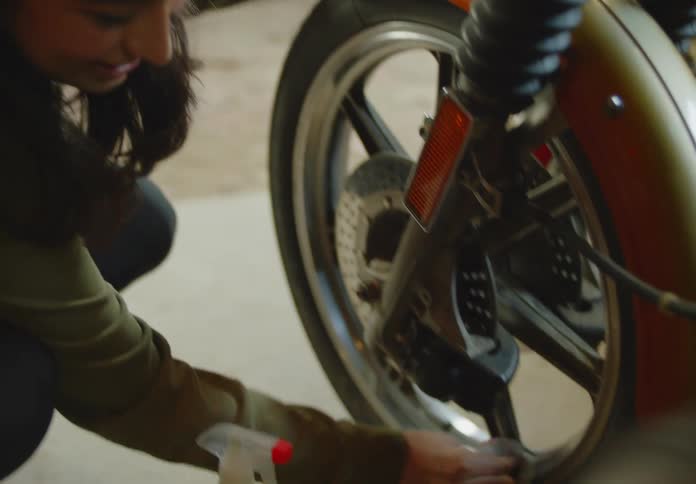 A young, ethnic woman with a cloth in hand chats with someone as she cleans and polishes the wheel of an orange motorbike.