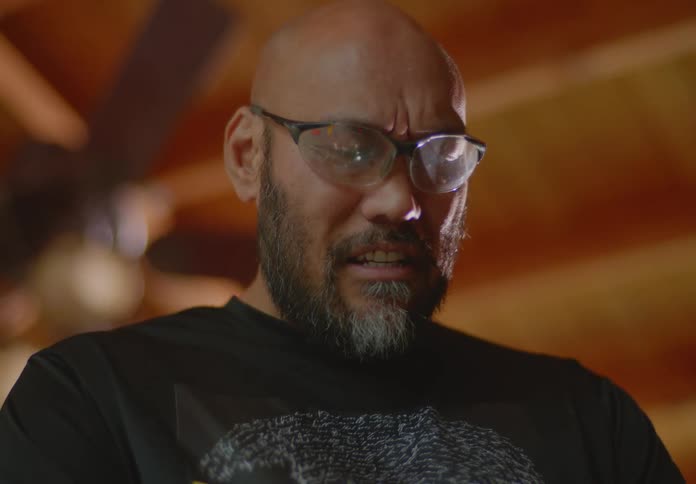 A tan, bearded man with bald head and safety glasses examines a metal pipe and blows on it to clean the edges as he talks with someone in the room