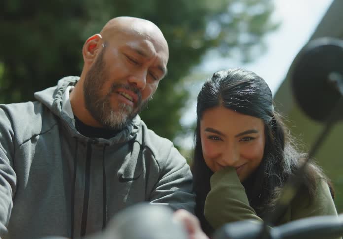 A tan and fit bald man with facial hair sits on his motorcycle and chats with a young, dark-haired woman standing next to him who is leaning in flirtatiously and showing interest.
