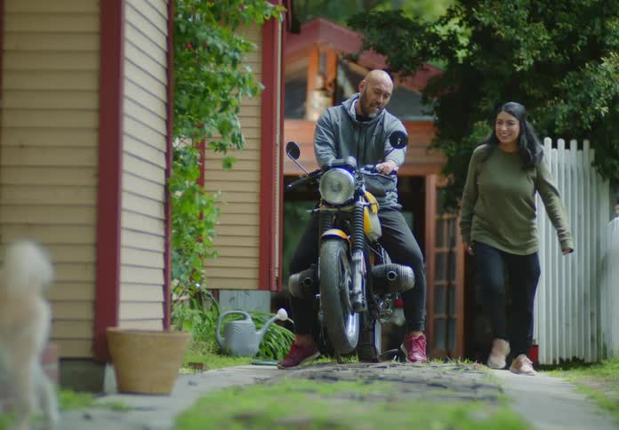A couple stands in their rural driveway discussing the motorbike the man is sitting on as their two dogs run by playing with each other.