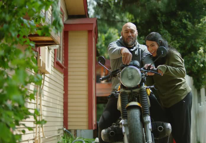 A bald, bearded man sits on a motorcycle fiddling with buttons as a dark-haired woman leans on him and the motorcycle and closes her eyes in a squint so he can surprise her with something as they laugh and flirt.