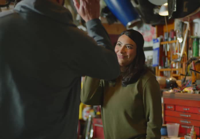 A dark-haired woman and a bald, bearded man give each other a high-five excitedly after installing a headlight on a motorcycle, and then the man turns and gleefully rubs his hands together.