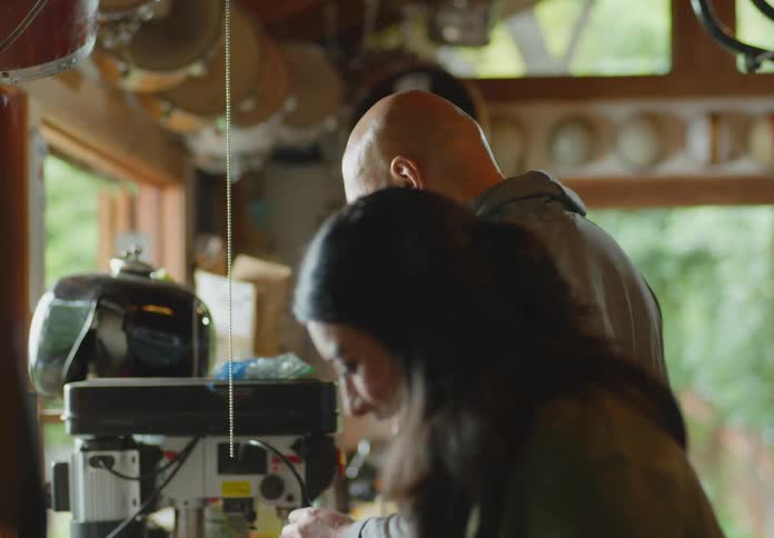 A man and a woman going through items on a garage workbench and the woman laughs after she finds an old ipod protective cover.