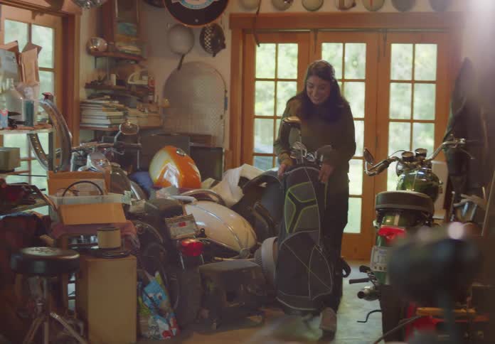 A young dark-haired woman carries a bag of golf clubs through a cluttered storage room and over to a man standing at a workbench.