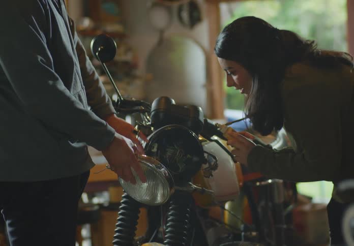 A dark-haired young woman applies glue to a motorcycle light casing as a man holds a headlight ready to install.