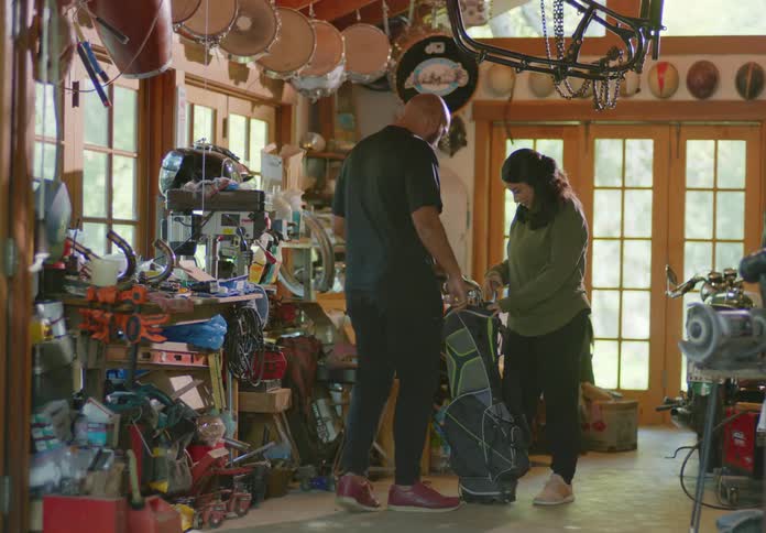 A young, brunette woman standing in a cluttered garage finds and drags a golf bag full of clubs over to a bald, bearded man and they each pull one out and examine it.