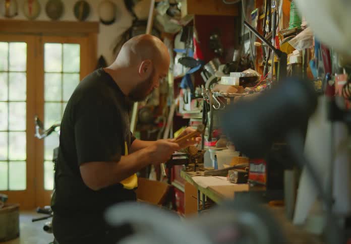 Side view through the clutter of a bald, bearded man at his workbench in a messy workshop working on a project with a hammer and chisel.