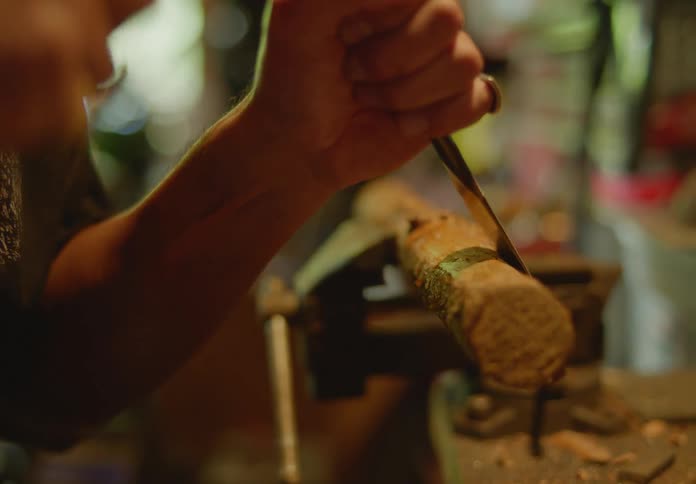 Hands of a carpenter using a hammer and chisel to remove bark shavings in a downward motion from a small log being held in a vice.