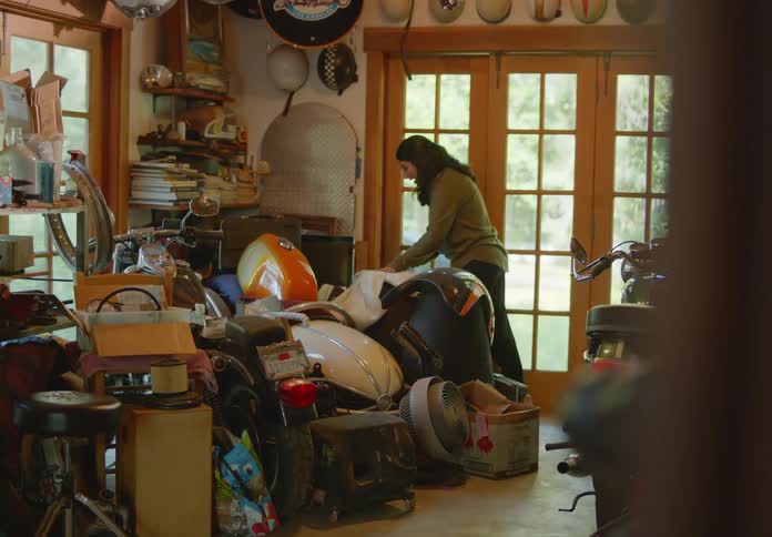A young woman meanders through a cluttered storage room with lots of natural light, finds an old case of golf clubs in the corner, pulls it out, holds it up, and talks with someone in the room about it.