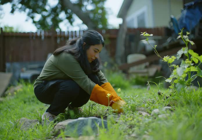 A woman kneels in front of a garden and yanks out a root from the ground