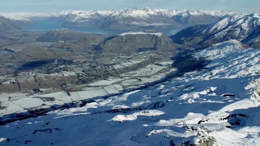 An aerial view of Coronet Peak mountain range that is partially covered in snow and Queenstown in distance