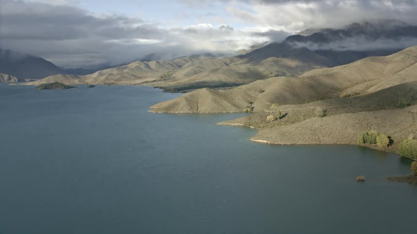 Flying across a  bay of water and in the sky there are several low hanging clouds and brown hills