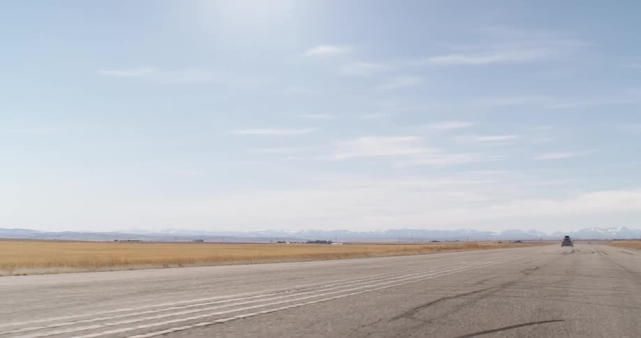 A car with a roof box is driving on a road through a flat landscape with mountains in the distance