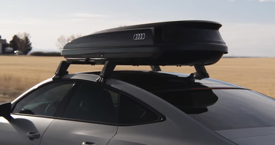 A grey audi car equipped with a black roof rack travels along a rural road adjacent to a farmhouse