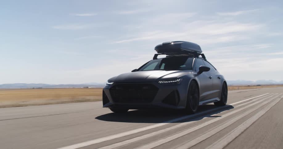 A car with a roof box is driving on a road through a flat landscape with mountains in the distance