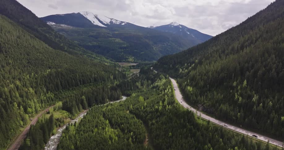 Aerial view of a winding road through a dense forest in the mountains with a river running parallel to it