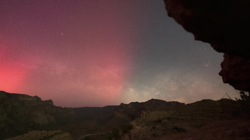 A gradient of pink and purple aurora in the night sky above a dark silhouette of mountainous terrain