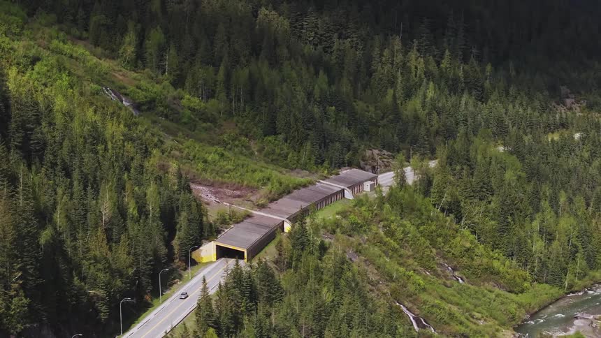 Vehicles drive on highway passing through long tunnel on forested mountain bordering river
