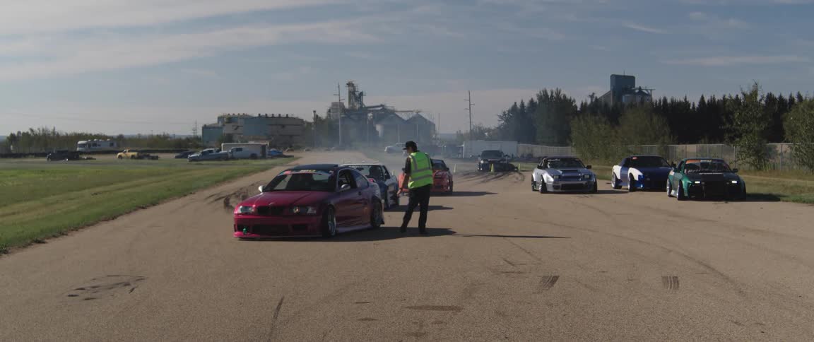 Group of cars on a race track with industrial structures in the background and a person in a green coat helping