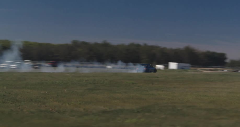 A blue car is doing donuts and emitting lots of smoke in a large field